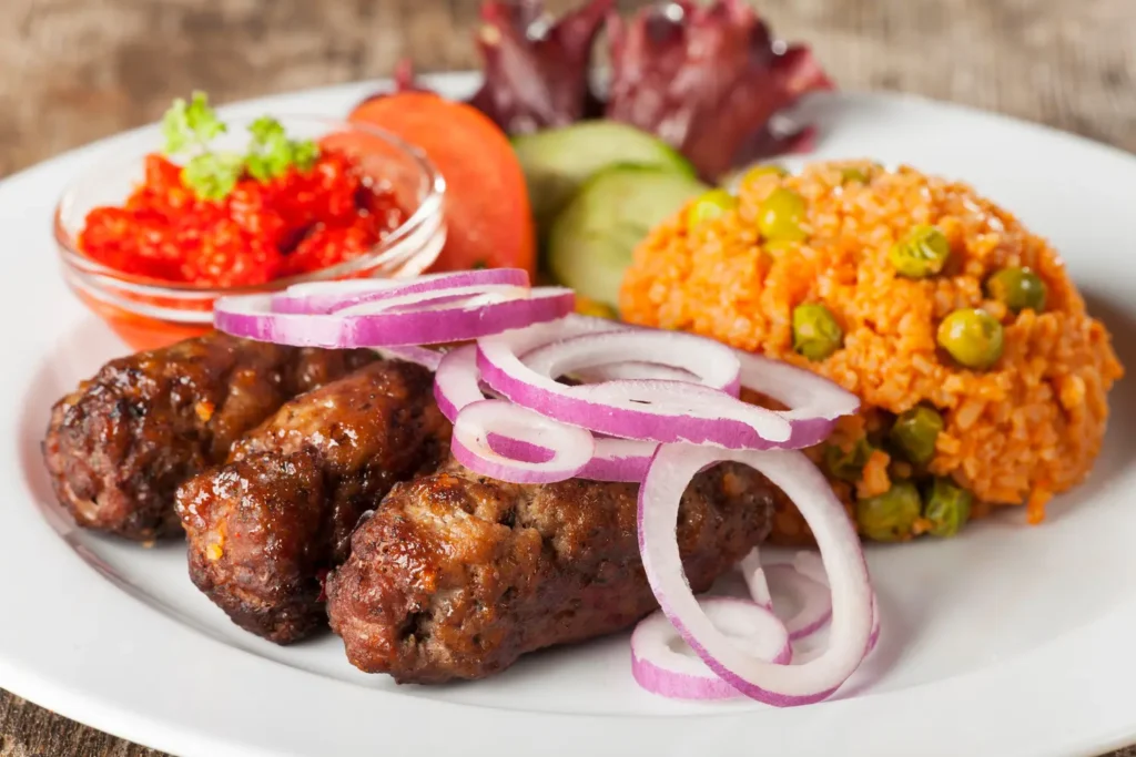 Drei gut gewürzte Cevapcici mit roten Zwiebeln, Reis und frischem Gemüse auf weißem Teller